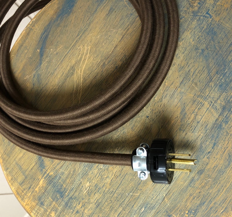 May include: Brown cloth-covered electrical cord with a black and silver plug. The cord is coiled on a wooden surface.