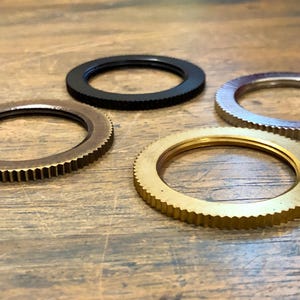 Op de afbeelding: Vier metalen ringen met een gegroefde buitenrand zijn gerangschikt op een houten oppervlak. De ringen zijn in verschillende kleuren: brons, zwart, zilver en goud. De ringen lijken componenten of onderdelen van een groter mechanisme te zijn.
