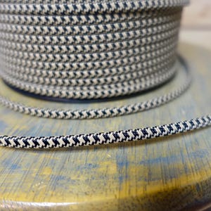 May include: A spool of navy blue and cream-colored rope with a repeating chevron pattern. A length of the same rope is unspooled and lies in front of the spool. The rope is on a light blue and yellow surface.
