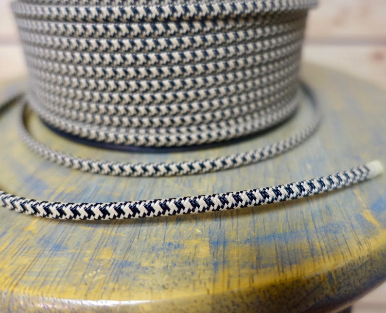 May include: A spool of navy blue and cream-colored rope with a repeating chevron pattern. A length of the same rope is unspooled and lies in front of the spool. The rope is on a light blue and yellow surface.