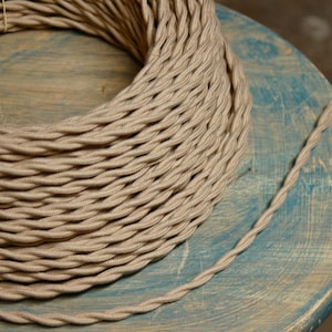 May include: A close-up of a reel of beige twisted electrical cable. The cable is made of fabric and has a subtle texture. The cable is coiled neatly on a wooden surface.