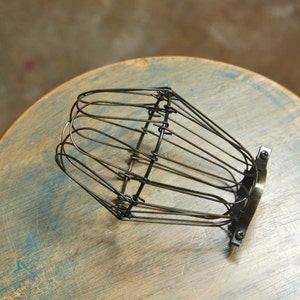 Op de afbeelding: Een zwarte metalen kooilampenkap met een draadframe ontwerp. De kooi is bevestigd aan een metalen basis met twee schroeven.