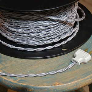 May include: A spool of silver braided electrical cord with a white plug attached. The cord is wound around a black plastic spool.
