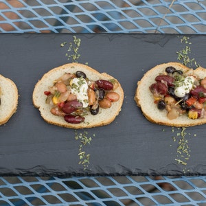 May include: Three slices of toasted bread topped with a colorful bean salad and a dollop of sour cream. The bread is arranged on a black slate serving tray.