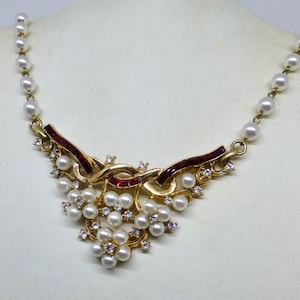 Op de afbeelding: Een goudkleurige ketting met een centrale hanger met imitatieparels, rode accenten en heldere kristallen. De ketting heeft aan elke kant een streng witte imitatieparels. De hanger heeft een bloemenontwerp.