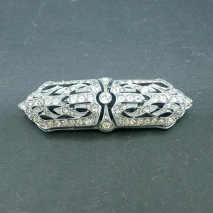 May include: A silver-toned brooch with a geometric design, featuring a central oval surrounded by clear rhinestones.