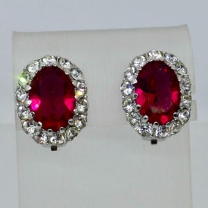以下が含まれることがあります： 大きな楕円形のルビーレッドの石が特徴のクリップオンイヤリングのペア。小さな透明なクリスタルが周囲を取り囲んでいます。イヤリングはシルバーカラーの金属製で、白いスタンドに展示されています。