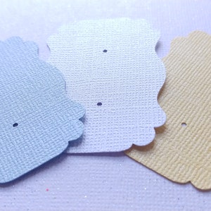Peut inclure: Trois cartons d'exposition de boucles d'oreilles aux couleurs pastel : bleu clair, blanc et jaune. Chaque carton a un bord festonné et deux petits trous pour les boucles d'oreilles. Les cartons ont une surface texturée sur fond blanc.