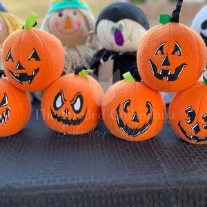 Könnte beinhalten: Sechs orangefarbene Plüschkürbisse mit verschiedenen gestickten Gesichtsausdrücken. Die Kürbisse sind in einem Halbkreis auf einer schwarzen Oberfläche angeordnet.