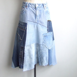 Può includere: Gonna di jeans color azzurro chiaro con design patchwork. La gonna presenta varie tonalità di denim blu, tra cui tasche e cuciture, creando un look asimmetrico unico. La gonna ha un orlo grezzo ed è esposta su un manichino.