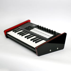 Puede incluir: Un controlador de teclado MIDI negro con teclas blancas, una cuadrícula de pads cuadrados retroiluminados y una fila de perillas giratorias. El dispositivo está montado en un soporte con paneles laterales negros brillantes y dos tornillos.
