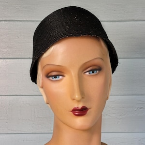 May include: A black, round hat with a textured surface. The hat is worn on a mannequin with a light skin tone, blonde hair, and red lipstick.