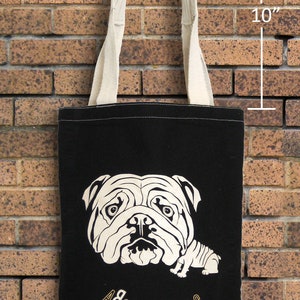 May include: Black tote bag with a cream-colored bulldog graphic and cream-colored handles. The bag is 10 inches in height. The text "ABSOLUTE DOG" is printed in gold. The bag is hanging on a brick wall.