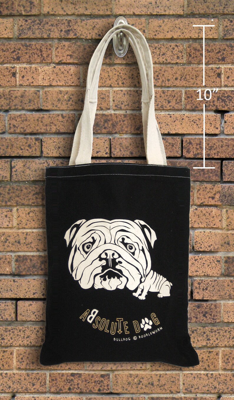 May include: Black tote bag with a cream-colored bulldog graphic and cream-colored handles. The bag is 10 inches in height. The text "ABSOLUTE DOG" is printed in gold. The bag is hanging on a brick wall.