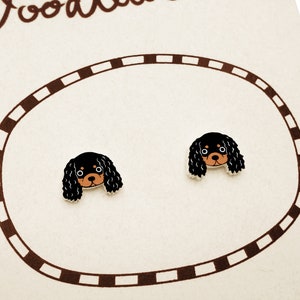 May include: A pair of stud earrings featuring a cartoon illustration of a black and tan Cavalier King Charles Spaniel dog head. The earrings are displayed on a light-colored card with a brown dashed oval border and partial text at the top.