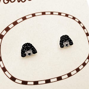 May include: A pair of poodle-shaped earrings. Each earring features a black poodle head with a white face and a detailed design. The earrings are set against a cream-colored background with a brown oval border.