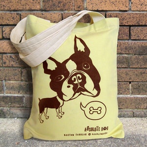 May include: A light yellow tote bag with a brown Boston Terrier graphic. The bag has a cream-colored strap and features the text "Absolute Dog" and "Boston Terrier @ Doodleworm". The dog graphic includes a speech bubble with a bone.