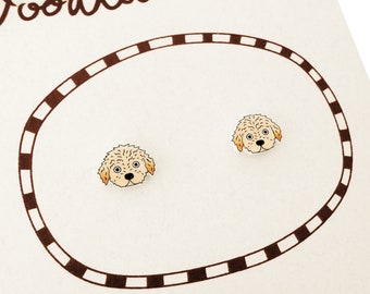 Può includere: Un paio di orecchini a lobo con illustrazioni di volti di cani dal pelo riccio color marrone chiaro, con naso nero e occhi grandi. Gli orecchini sono esposti su un cartoncino bianco con un bordo ovale a tratti marroni e bianchi.