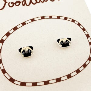 May include: A pair of stud earrings featuring a cartoon pug dog face design with tan and black coloring. The earrings are displayed on a cream-colored card with a brown circular border and the partial text Doodl at the top.