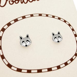 May include: A pair of raccoon-faced stud earrings. Each earring features a gray and white raccoon face with black accents. The earrings are displayed on a cream-colored card with a brown oval border.