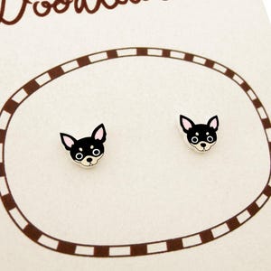 Puede incluir: Un par de pendientes de botón de Chihuahua. Cada pendiente presenta la cabeza de un Chihuahua negro y blanco con orejas rosas. Se presentan en una tarjeta crema con un borde ovalado marrón.