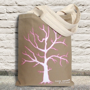 May include: A tan tote bag with cream-colored handles and a white tree design with pink accents. The words "love nature" and the copyright are printed at the bottom of the bag. The bag is made of canvas.