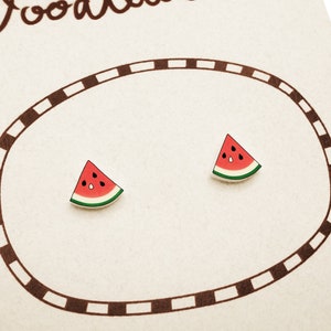 Könnte beinhalten: Ein Paar Ohrstecker in Form von Wassermelonenscheiben mit rotem Fruchtfleisch, weißer Schale, grünem Rand und schwarzen Kernen. Die Ohrstecker sind auf einer hellen Karte mit einem braunen, gestrichelten ovalen Rahmen und Textfragmenten befestigt.