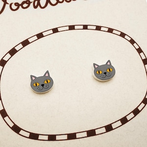 May include: A pair of gray cat-shaped stud earrings with yellow eyes and a smiling face. The earrings are set on a cream-colored card with a brown oval border. The word "Doodles" is visible in the upper left corner.
