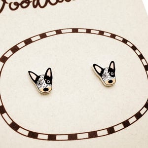 May include: A pair of dog-shaped stud earrings. Each earring features a white, black, and tan dog face design. The earrings are displayed on a cream-colored card with a brown oval border.