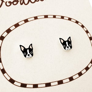 May include: A pair of stud earrings featuring a Boston Terrier dog design. The earrings are black and white, with a detailed illustration of the dog's face. They are displayed on a cream-colored card with a brown oval border.