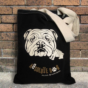 May include: Black tote bag featuring a cream-colored illustration of a bulldog's face and body. The bag has a cream-colored strap and the words "ABSOLUTE DOG" and "BULLDOG @ DOODLEWORMS" printed in gold.