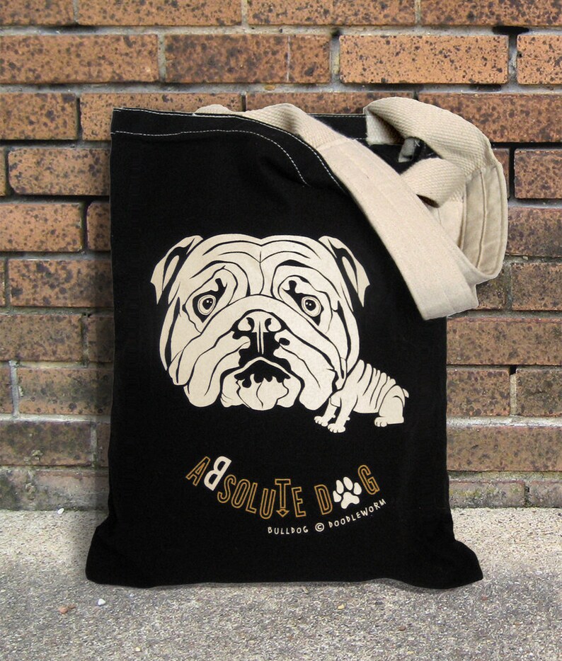 May include: Black tote bag featuring a cream-colored illustration of a bulldog's face and body. The bag has a cream-colored strap and the words "ABSOLUTE DOG" and "BULLDOG @ DOODLEWORMS" printed in gold.