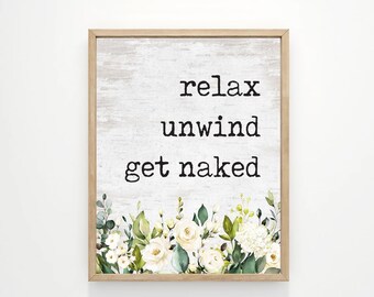 May include: A framed wall art print with the words "relax unwind get naked" in black font on a white and gray background. The bottom of the print features a floral design with white roses and green leaves. The frame is a light brown color.