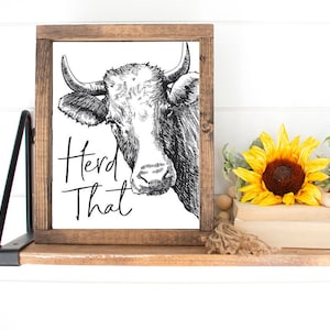 May include: A black and white illustration of a cow's head with the text "Heard That" on a white background. The illustration is framed in a brown wooden frame.