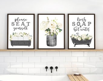 May include: Three framed bathroom prints with white flowers and text. The first print says "please seat yourself" with a drawing of a toilet paper holder. The second print has a mason jar with white flowers and the text "Hello Sweet Cheeks". The third print has a drawing of a bathtub with white flowers and the text "fresh soap & hot water".