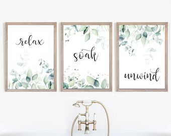 May include: Three framed prints with watercolor eucalyptus leaves and the words "relax", "soak", and "unwind" on a white background. The prints are hanging on a white wall above a bathtub.