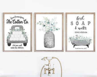 May include: Three framed prints with a farmhouse theme. The first print features a vintage truck delivering toilet paper with the text "Finest Premium Quality The Cotton Co. 25¢ per roll same day delivery". The second print features a mason jar filled with eucalyptus leaves and the text "The Handmade Soap Co.". The third print features a bathtub with eucalyptus leaves and the text "Fresh SOAP & water FIVE CENTS hand towels extra".