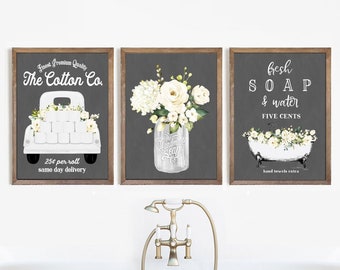 May include: Three framed prints with a farmhouse theme. The first print features a white pickup truck with a sign that reads "Finest Premium Quality The Cotton Co. 25¢ per roll same day delivery". The second print features a mason jar filled with white flowers and a sign that reads "The Handmade Soap Co.". The third print features a white bathtub with a sign that reads "Fresh SOAP & water FIVE CENTS hand towels extra".