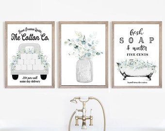 May include: Three framed prints with a bathroom theme. The left print features a white pickup truck with a stack of toilet paper rolls and the text "Finest Premium Quality The Cotton Co. 25¢ per roll same day delivery". The middle print features a mason jar with a bouquet of eucalyptus and the text "The Handmade Soap". The right print features a bathtub with eucalyptus and the text "Fresh SOAP & water FIVE CENTS hand towels extra".