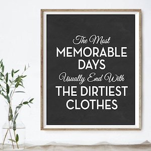 Op de afbeelding: Zwart-wit typografieprint op een zwarte achtergrond met een houten frame. De tekst luidt "The Most Memorable Days Usually End With The Dirtiest Clothes."