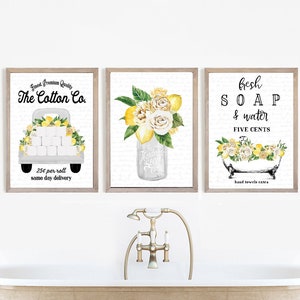 May include: Three framed prints with a lemon and floral theme. The first print features a white truck with toilet paper rolls and lemons, with the text "Finest Premium Quality The Cotton Co. 25c per roll same day delivery". The second print features a mason jar with lemons and flowers, with the text "The Handmade Soap Co.". The third print features a bathtub with lemons and flowers, with the text "fresh SOAP & water FIVE CENTS hand towels extra".