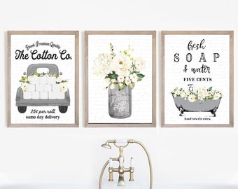May include: Three framed bathroom art prints. The prints feature a gray truck filled with toilet paper and flowers, a mason jar with flowers, and a bathtub with flowers. The text includes "The Cotton Co.", "Handmade Soap", and "Fresh Soap & Water".
