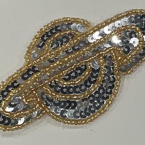 Può includere: Applicazione decorativa con un design a spirale. L'applicazione è impreziosita da paillettes argentate e perline color oro. I bordi sono delineati con perline dorate, creando un accessorio strutturato e accattivante.