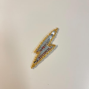 Könnte beinhalten: Ein dekorativer Aufnäher in Form eines Blitzes. Der Aufnäher ist mit silbernen Pailletten verziert und mit goldenen Perlen umrandet. Die Mitte des Blitzes hat eine Mischung aus silbernen und roten Pailletten. Dieses Accessoire kann auf Kleidung oder Accessoires genäht oder aufgebügelt werden.