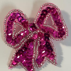 May include: A pink sequined bow with silver beads around the edges. The bow is made of a sheer fabric with sequins sewn on top.