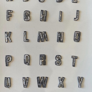 May include: A set of 26 silver and black iron-on alphabet letters. Each letter is decorated with silver sequins.