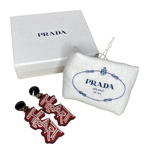 May include: A pair of red heart-shaped earrings with black accents, packaged in a white box with the Prada logo. The box is also included in the image.