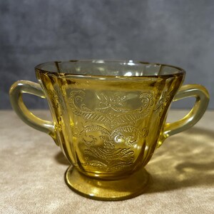 May include: Amber-colored glass sugar bowl with two handles and a pedestal base. The bowl features an embossed floral design on the front and back. The glass has a slightly textured appearance.