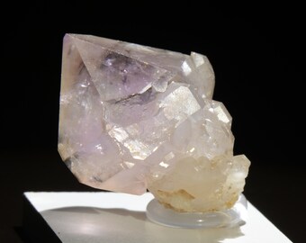 May include: A translucent, light purple quartz crystal cluster. The main crystal is a large, rectangular shape with a pointed top. Smaller, clear crystals are clustered at the base. The crystal sits on a clear, circular stand against a black background.