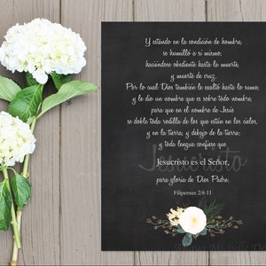 May include: A black chalkboard-style print with white text, featuring a Bible verse in Spanish, and a floral design at the bottom. Two white hydrangea blooms and green leaves are on the left side, set against a wooden background.